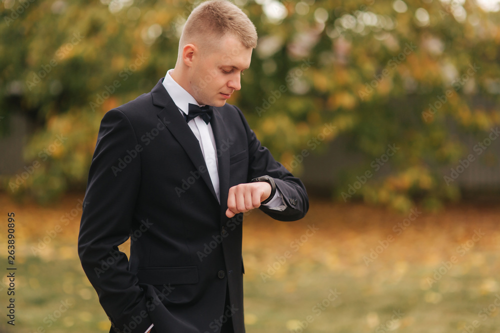 Handsome man in suit look at the watch outside, autumn. Man in black suit with beautiful tie bow Handsome man in suit look at the watch outside, autumn. Man in black suit with beautiful tie bow