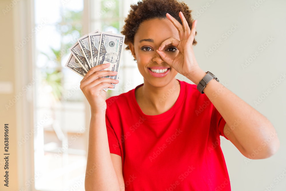 Young african american woman holding bank notes of fifty dollars with happy face smiling doing ok sign with hand on eye looking through fingers Young african american woman holding bank notes of fifty dollars with happy face smiling doing ok sign with hand on eye looking through fingers