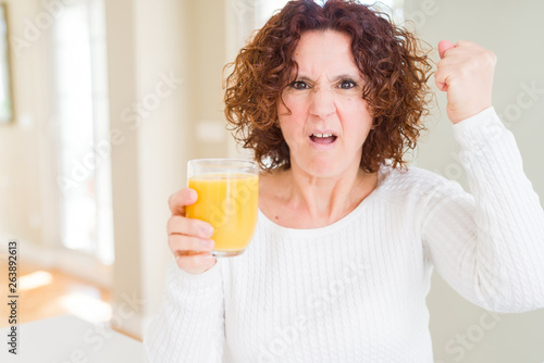 Senior woman driking a glass of fresh orange juice annoyed and frustrated shouting with anger, crazy and yelling with raised hand, anger concept