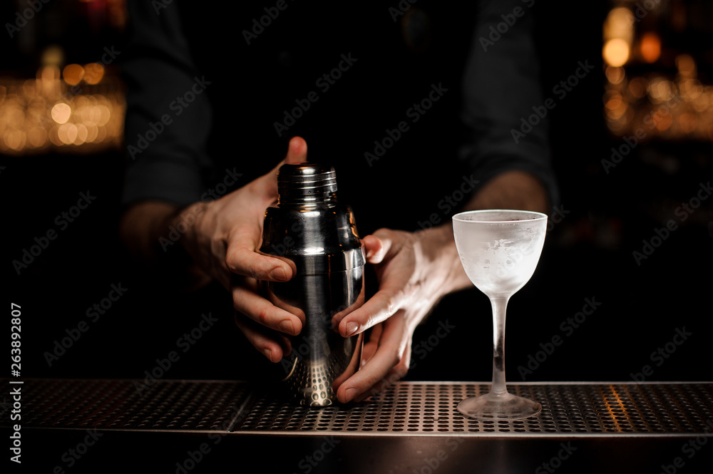 Bartender holds shaker with alcohol cocktail glass nearby
