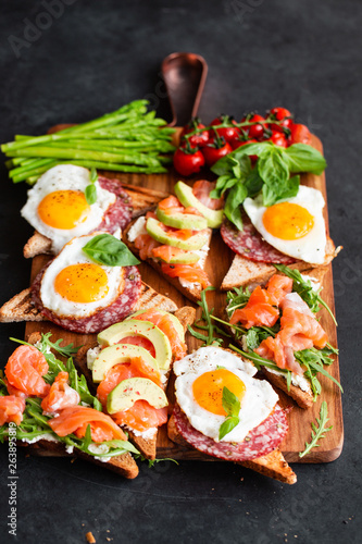Huge healthy breakfast with sandwiches with scrambled eggs, sausage, salmon, arugula, curd cheese, avocado on a wooden board, sun-dried tomatoes, asparagus, radishes, greens on black table top view