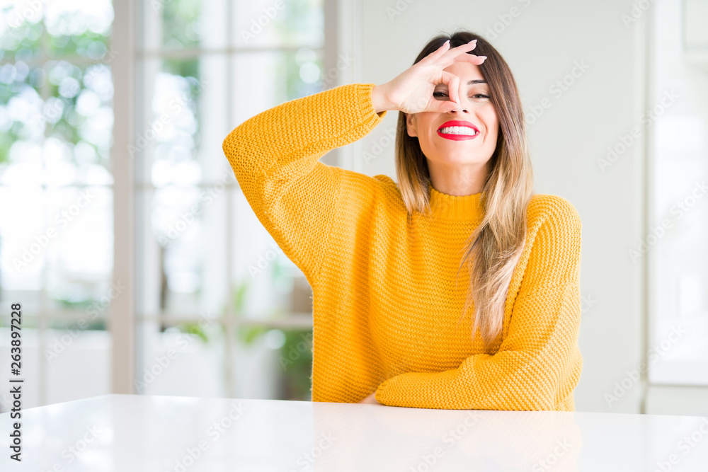 Young beautiful woman wearing winter sweater at home doing ok gesture with hand smiling, eye looking through fingers with happy face. Young beautiful woman wearing winter sweater at home doing ok gesture with hand smiling, eye looking through fingers with happy face.