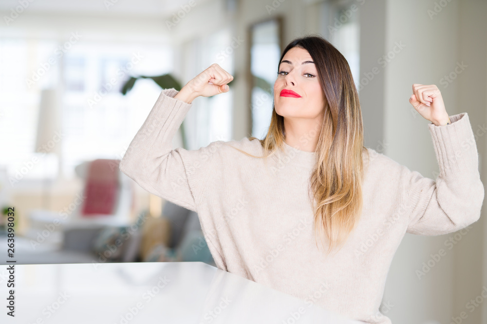 Young beautiful woman wearing winter sweater at home showing arms muscles smiling proud. Fitness concept. Young beautiful woman wearing winter sweater at home showing arms muscles smiling proud. Fitness concept.
