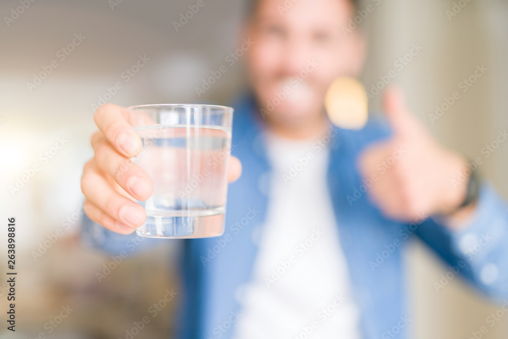 Young handsome man drinking a glass of water at home happy with big smile doing ok sign, thumb up with fingers, excellent sign Young handsome man drinking a glass of water at home happy with big smile doing ok sign, thumb up with fingers, excellent sign