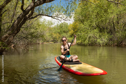 A young woman wearing sunglasses and a swimming suit sits on a paddle board paddling down Quail creek in Southern Utah with willow trees lining the banks on either side. 