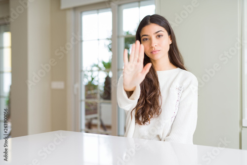 Young beautiful woman at home on white table doing stop sing with palm of the hand. Warning expression with negative and serious gesture on the face.