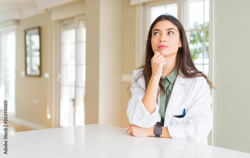 Young woman wearing medical coat at the clinic as therapist or doctor with hand on chin thinking about question, pensive expression. Smiling with thoughtful face. Doubt concept. Young woman wearing medical coat at the clinic as therapist or doctor with hand on chin thinking about question, pensive expression. Smiling with thoughtful face. Doubt concept.