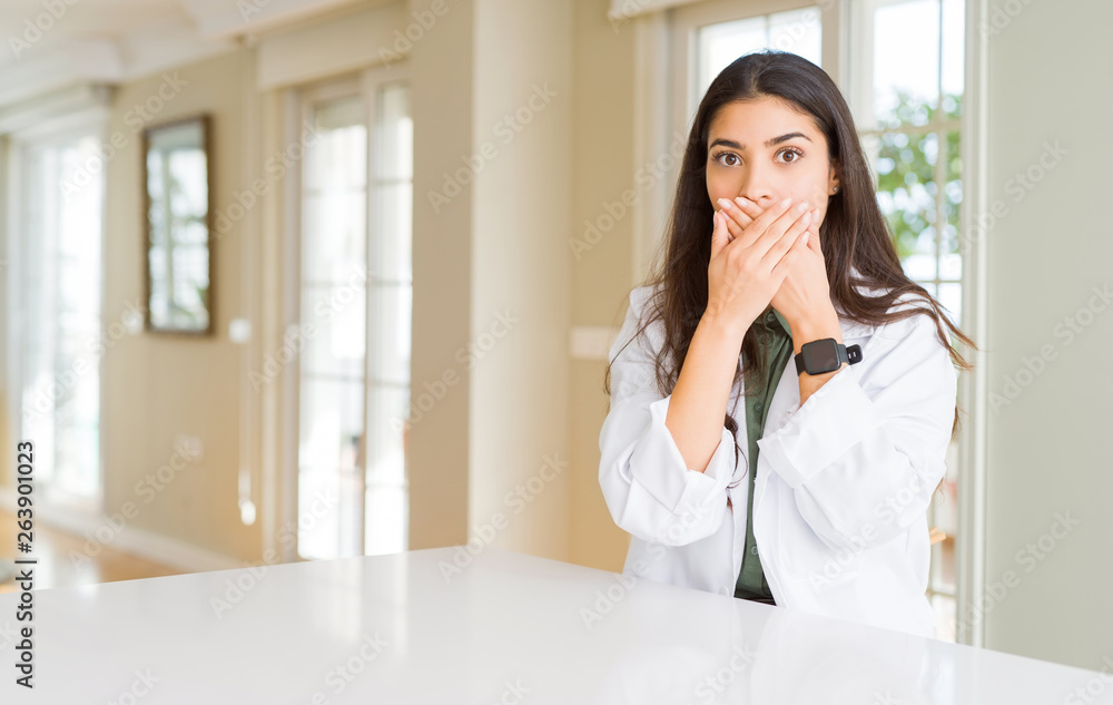 Young woman wearing medical coat at the clinic as therapist or doctor shocked covering mouth with hands for mistake. Secret concept. Young woman wearing medical coat at the clinic as therapist or doctor shocked covering mouth with hands for mistake. Secret concept.