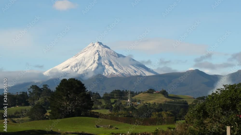 Snow capped mountain towers above green pasture in Taranaki, New Zealand on a sunny morning, TIME LAPSE, ZOOM. Snow capped mountain towers above green pasture in Taranaki, New Zealand on a sunny morning, TIME LAPSE, ZOOM.