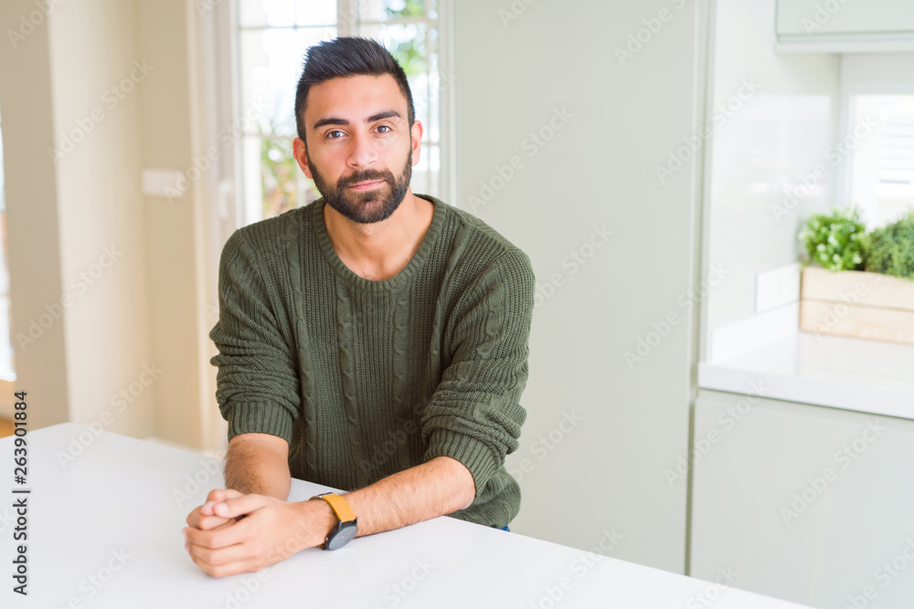 Handsome hispanic man wearing casual sweater at home with serious expression on face. Simple and natural looking at the camera. Handsome hispanic man wearing casual sweater at home with serious expression on face. Simple and natural looking at the camera.