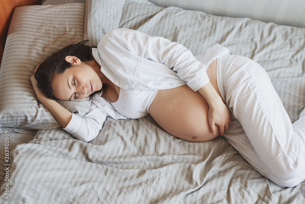 Young pregnant woman lying on bed hugging her tummy, healthy pregnancy - obrazy, fototapety, plakaty 