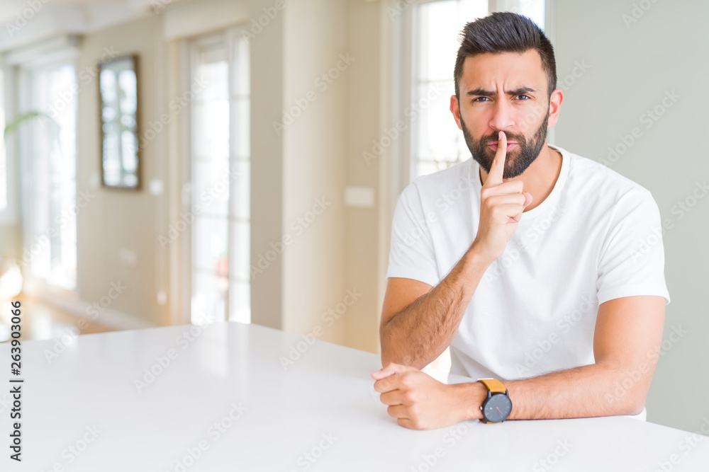 Handsome hispanic man casual white t-shirt at home asking to be quiet with finger on lips. Silence and secret concept.