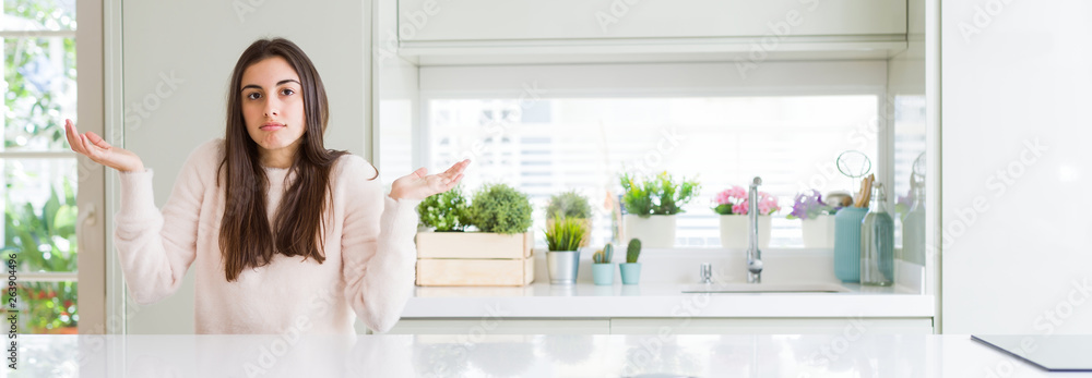 Wide angle picture of beautiful young woman sitting on white table at home clueless and confused expression with arms and hands raised. Doubt concept. Wide angle picture of beautiful young woman sitting on white table at home clueless and confused expression with arms and hands raised. Doubt concept.