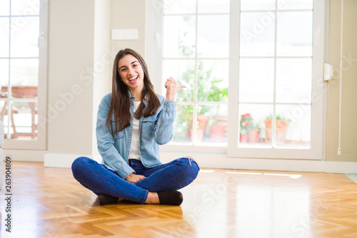 Beautiful young woman sitting on the floor at home smiling with happy face looking and pointing to the side with thumb up.