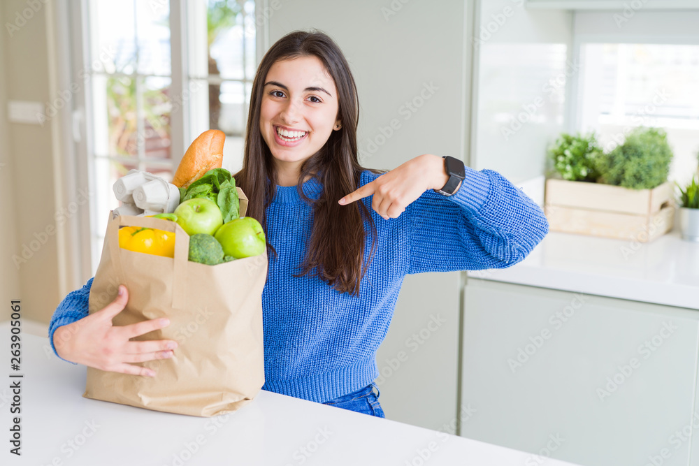 Beautiful young woman holding paper bag full of healthy groceries with surprise face pointing finger to himself Beautiful young woman holding paper bag full of healthy groceries with surprise face pointing finger to himself