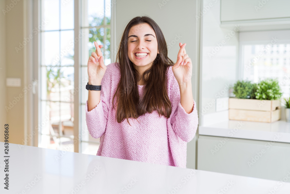 Beautiful young woman wearing pink sweater smiling crossing fingers with hope and eyes closed. Luck and superstitious concept.