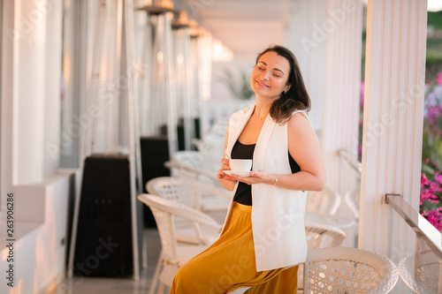 Stylish american woman at yellow dreess posed at cafe with cup of cappuccino at hands.