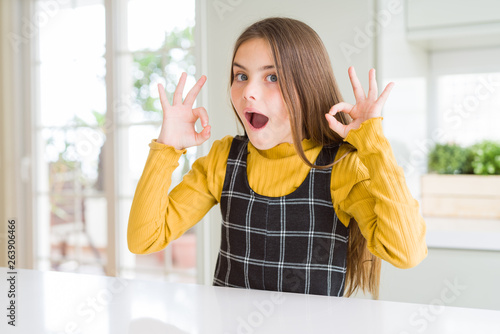 Young beautiful blonde kid girl wearing casual yellow sweater at home looking surprised and shocked doing ok approval symbol with fingers. Crazy expression