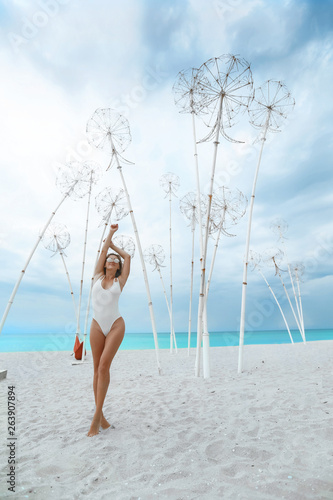 Outdoor shot of pleased young female with tanned skin, slim body, wears red bikini and sunglasses, poses against wonderful ocean view, blue sky, enjoys rest in resort place. People and recreation