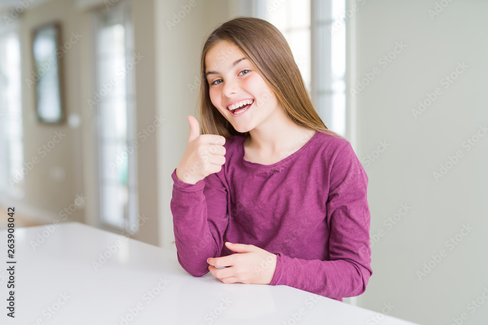 Beautiful young girl kid on white table doing happy thumbs up gesture with hand. Approving expression looking at the camera with showing success. Beautiful young girl kid on white table doing happy thumbs up gesture with hand. Approving expression looking at the camera with showing success.