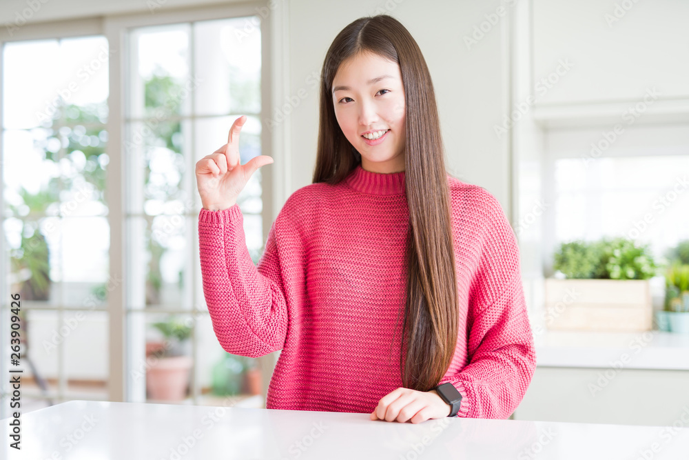 Beautiful Asian woman wearing pink sweater on white table smiling and confident gesturing with hand doing size sign with fingers while looking and the camera. Measure concept. Beautiful Asian woman wearing pink sweater on white table smiling and confident gesturing with hand doing size sign with fingers while looking and the camera. Measure concept.