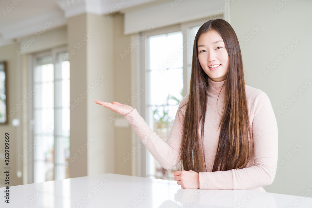 Beautiful Asian woman wearing casual sweater on white table smiling cheerful presenting and pointing with palm of hand looking at the camera. Beautiful Asian woman wearing casual sweater on white table smiling cheerful presenting and pointing with palm of hand looking at the camera.