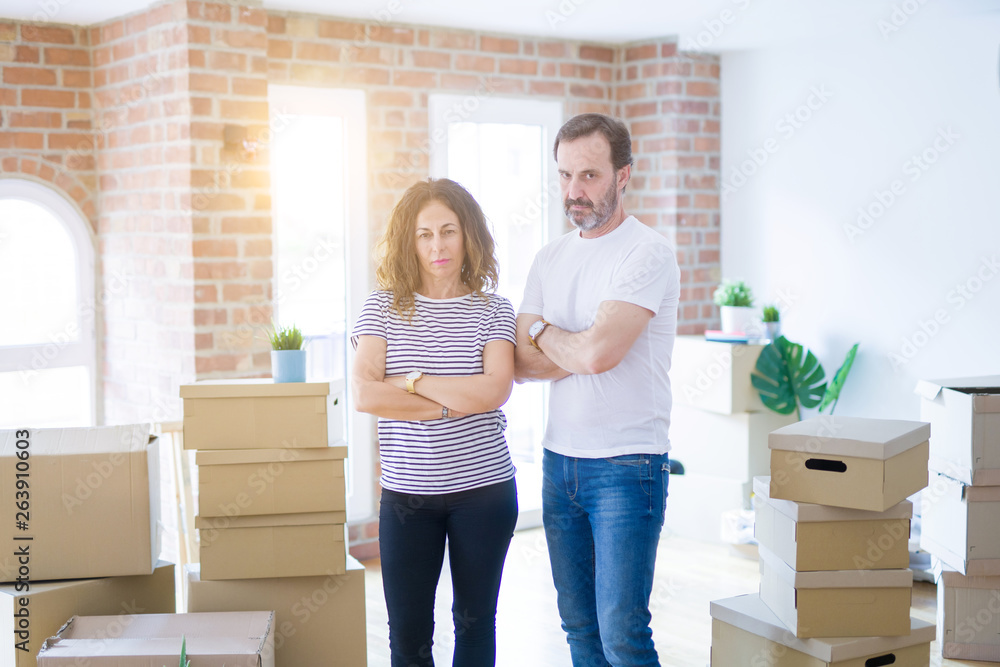 Middle age senior couple moving to a new home with boxes around skeptic and nervous, disapproving expression on face with crossed arms. Negative person. Middle age senior couple moving to a new home with boxes around skeptic and nervous, disapproving expression on face with crossed arms. Negative person.