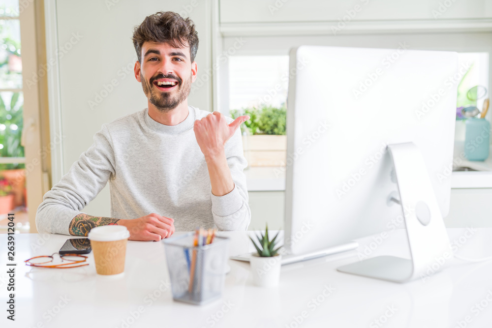 Young man working using computer smiling with happy face looking and pointing to the side with thumb up. Young man working using computer smiling with happy face looking and pointing to the side with thumb up.