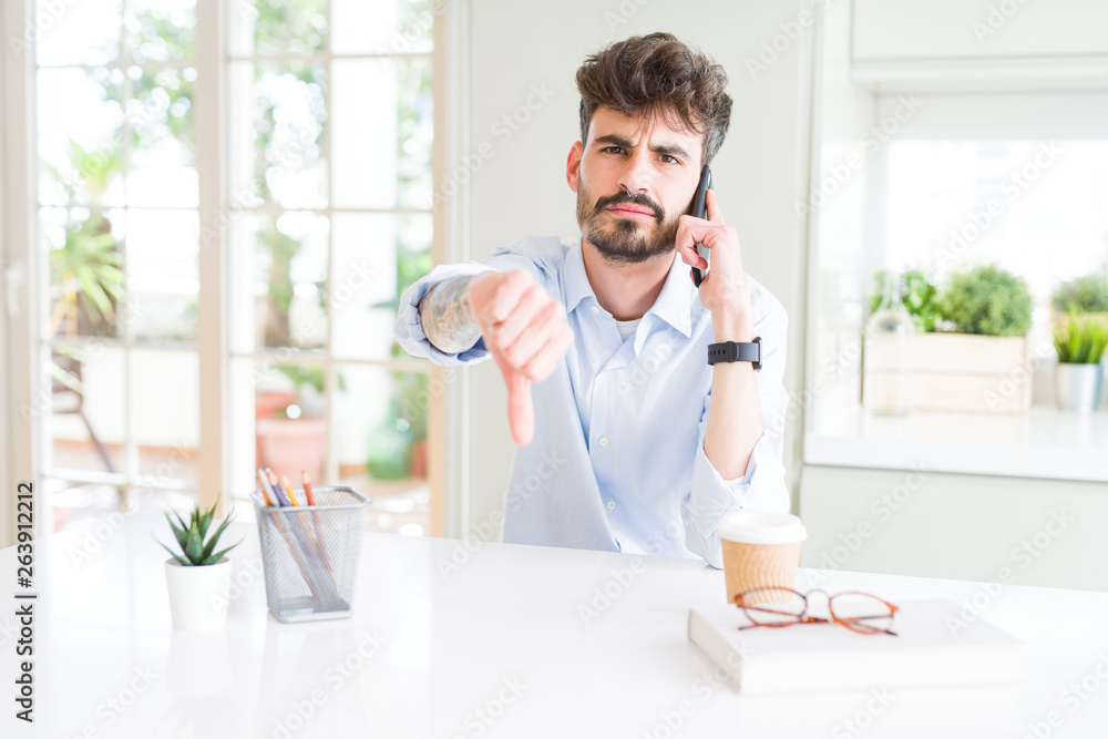 Young business man talking on smartphone with angry face, negative sign showing dislike with thumbs down, rejection concept