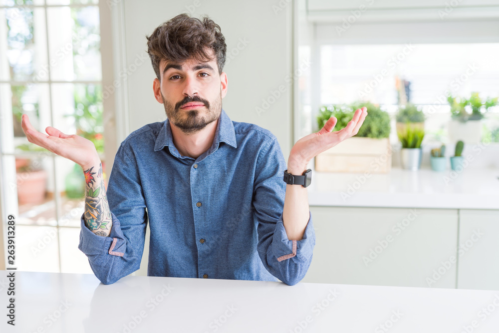 Young man wearing casual shirt sitting on white table clueless and confused expression with arms and hands raised. Doubt concept. Young man wearing casual shirt sitting on white table clueless and confused expression with arms and hands raised. Doubt concept.