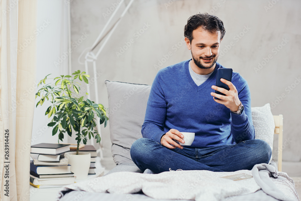 Attractive smiling arab man dressed casual sitting on bed in bedroom, drinking coffee and using smart phone for texting. Attractive smiling arab man dressed casual sitting on bed in bedroom, drinking coffee and using smart phone for texting.