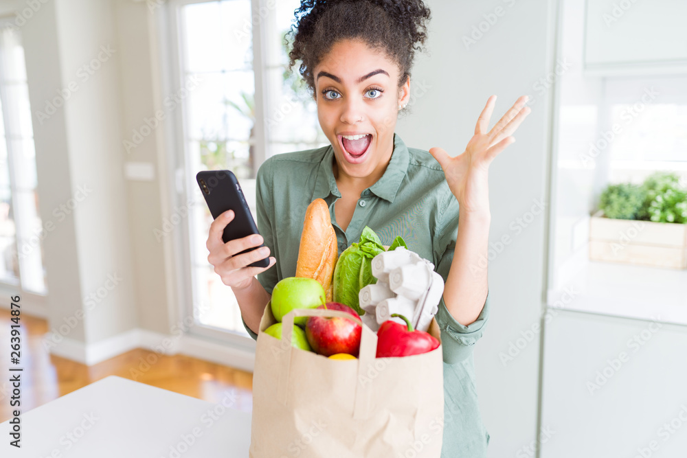 Young african american girl holding paper bag of groceries and using smartphone very happy and excited, winner expression celebrating victory screaming with big smile and raised hands Young african american girl holding paper bag of groceries and using smartphone very happy and excited, winner expression celebrating victory screaming with big smile and raised hands