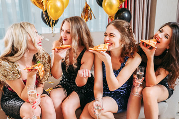 Girls party. Young ladies sitting in room decorated with balloons, eating pizza, chatting, smiling, having fun together.