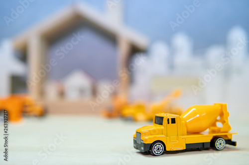 Miniature yellow concrete mixer truck model on wooden table with blurred house frame and city in the background. Warehouse logistics business or construction industry concept