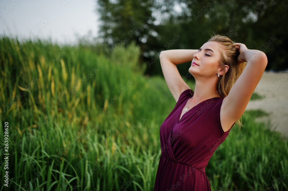 Blonde sensual woman in red marsala dress posing against lake with reeds. Blonde sensual woman in red marsala dress posing against lake with reeds.