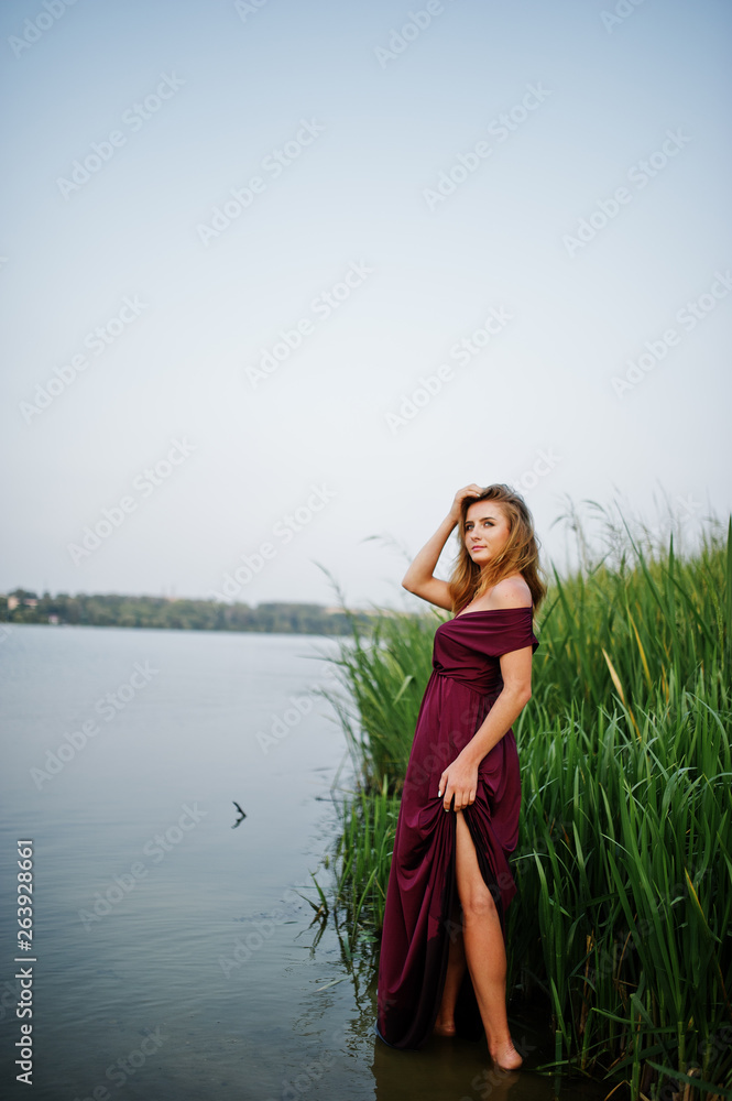 Obraz premium Blonde sensual woman in red marsala dress standing in water of lake with reeds.