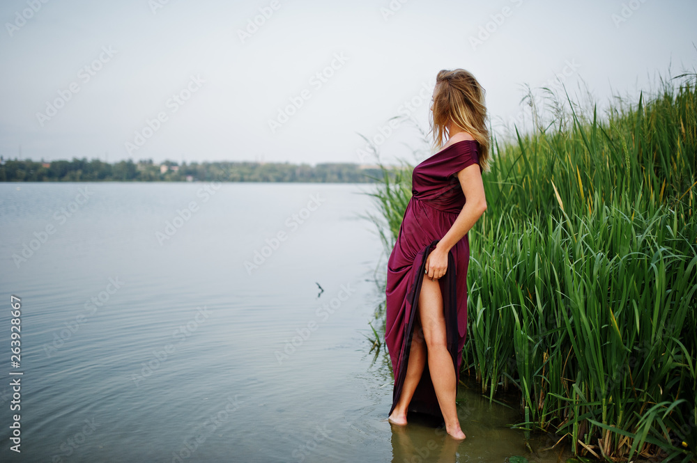 Fototapeta premium Blonde sensual woman in red marsala dress standing in water of lake with reeds.