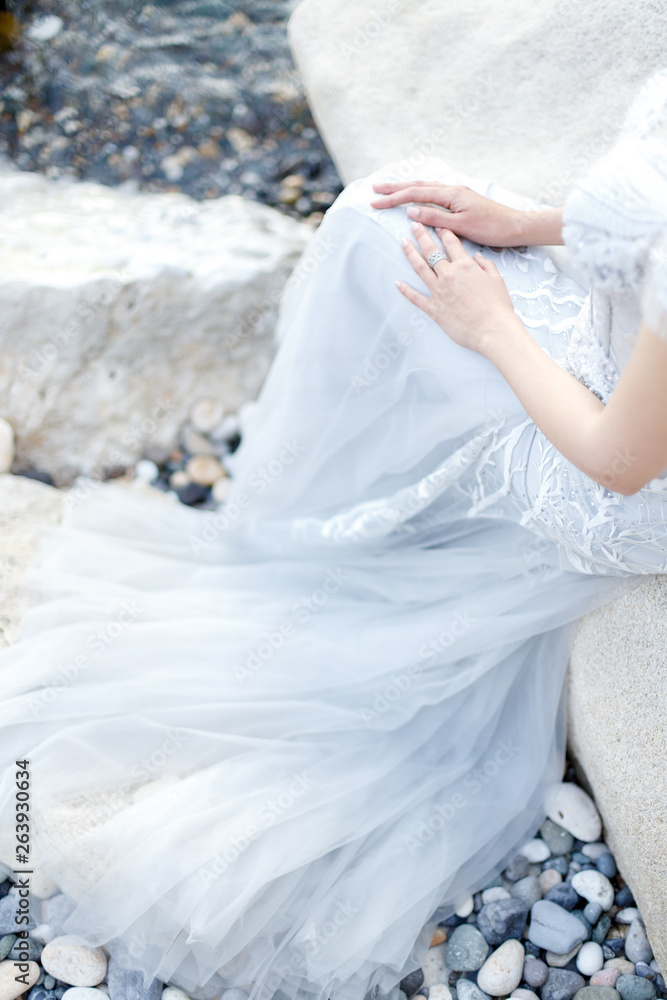 Woman legs, the hem of the dress, details. Bride in long light grey chiffon wedding dress embroidered with beads sitting on a rocky beach near the sea. No face. Woman legs, the hem of the dress, details. Bride in long light grey chiffon wedding dress embroidered with beads sitting on a rocky beach near the sea. No face.