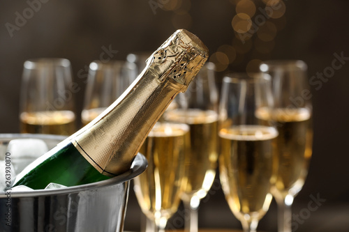 Bottle of champagne in bucket with ice and glasses on blurred background, closeup. Space for text