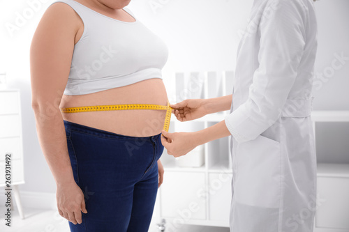 Doctor measuring waist of overweight woman in clinic, closeup photo