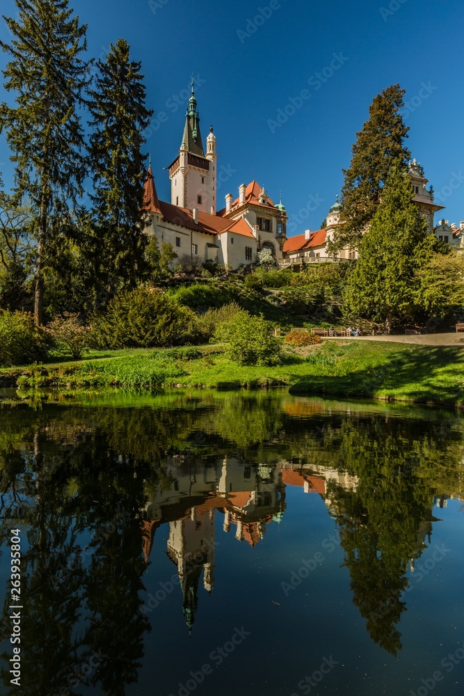 Pruhonice, Czech Republic - April 22 2019: Scenic landscape of famous romantic Pruhonice castle, standing on green hill in park, sunny spring day, blue sky, trees, reflection in water, vertical image