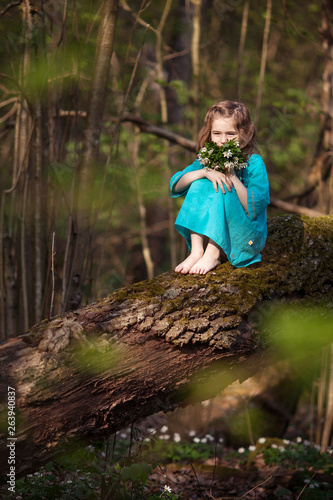 Fototapeta Naklejka Na Ścianę i Meble -  Beautiful little girl in a blue dress walking in the spring wood. Pretty  girl sitting on the tumbled-down tree. Kid playing outdoors in summer. Easter time.