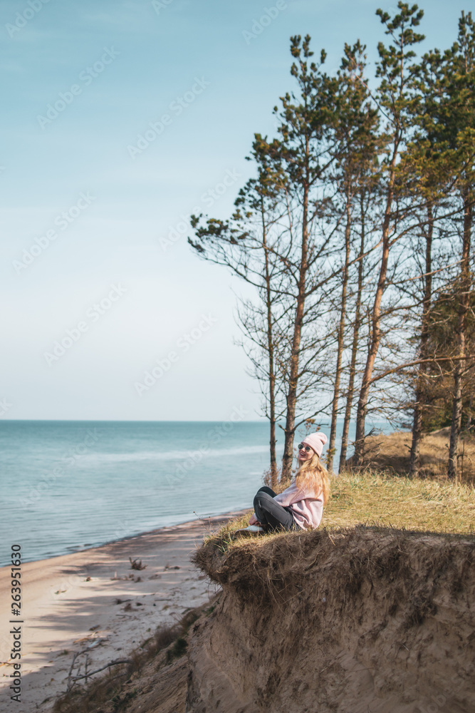 Obraz premium Young adult woman in pink hat sitting alone on the bluffs, looking of sea, freedom concept, peaceful atmosphere, meditation, copy space, Latvia, Jurkalne