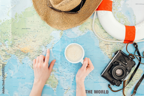 Cropped view of woman with cup of cappuccino, film camera, sunglasses and straw hat pointing with finger on world map