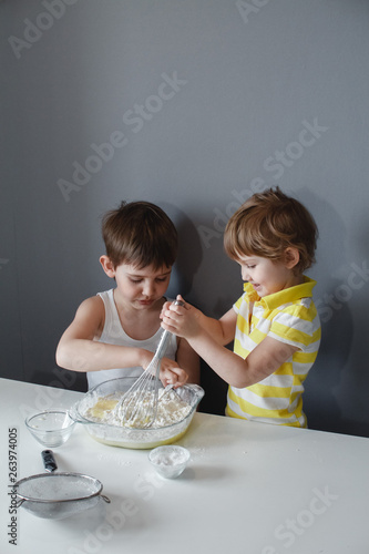 Two cheerful little children make shortbread. Adds flour to the dough. Sitting at a white table on a gray background. Preparation of desserts.