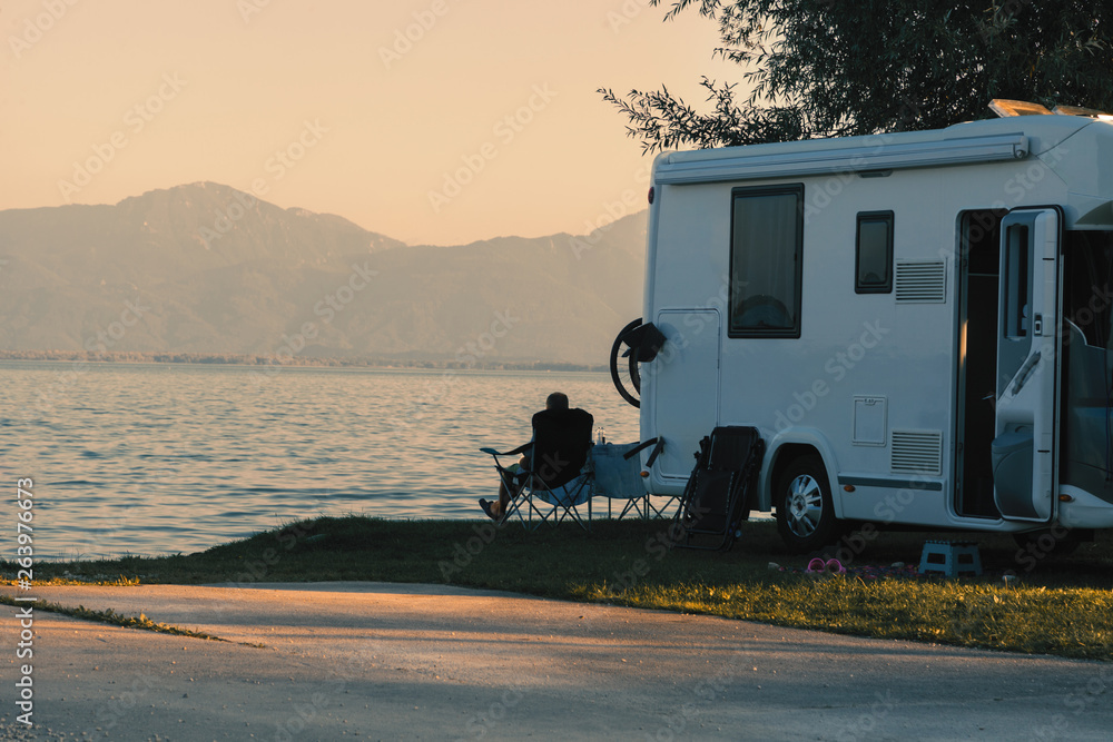 Man sitting near his caravan observes the sunset close the lake - obrazy, fototapety, plakaty 