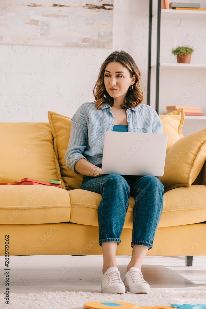 Attractive freelancer sitting on yellow sofa and using laptop Attractive freelancer sitting on yellow sofa and using laptop
