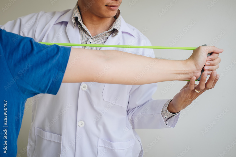 Doctor physiotherapist assisting a male patient while giving exercising treatment on stretching his arm with exercise band in the clinic, Rehabilitation physiotherapy concept