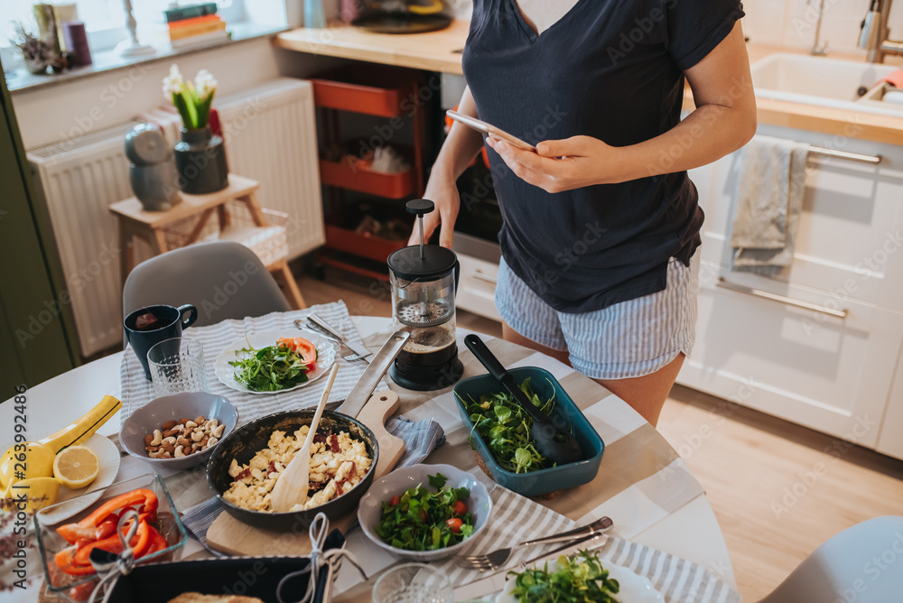 Breakfast table and woman holding smartphone in one hand navigating on social media and holding a big coffee filter glass mug with other hand, poring coffee. Breakfast table and woman holding smartphone in one hand navigating on social media and holding a big coffee filter glass mug with other hand, poring coffee.