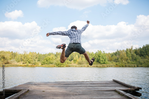 Photo from back of young jumping man on wooden bridge by river photo